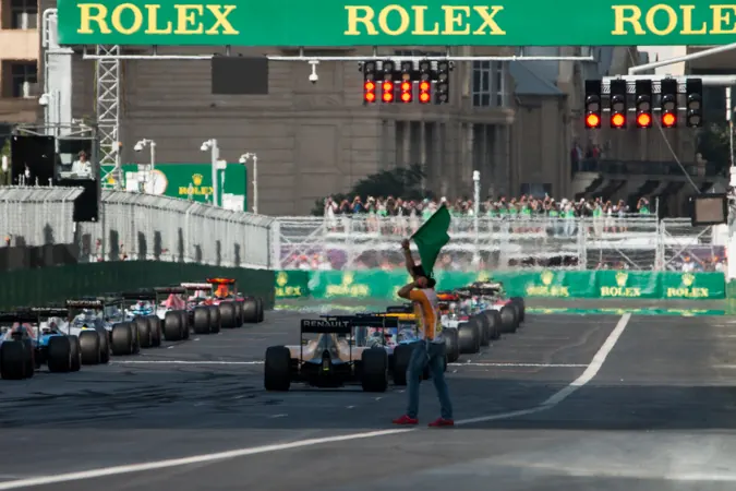 The grid lined up on the start/finish straight in Baku under the start lights, with city buildings behind the grandstand
